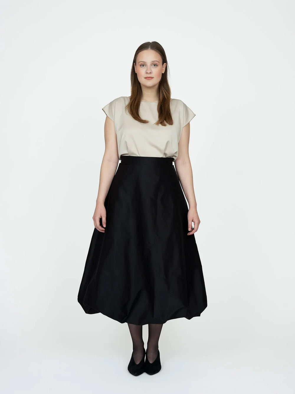 Woman wearing the Balloon Skirt sewing pattern from The Assembly Line on The Fold Line. A skirt pattern made in silk or wool fabric, featuring a flared silhouette, straight waistband, concealed zip closure, side seam pockets, and midi length with a balloon effect.