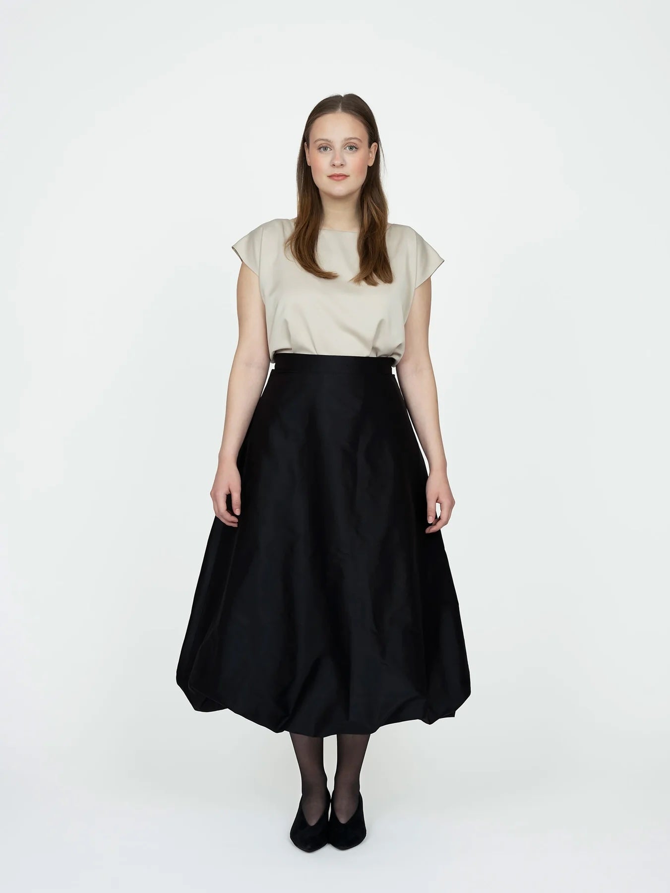 Woman wearing the Balloon Skirt sewing pattern from The Assembly Line on The Fold Line. A skirt pattern made in silk or wool fabric, featuring a flared silhouette, straight waistband, concealed zip closure, side seam pockets, and midi length with a balloon effect.