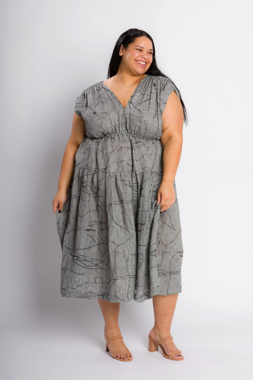 Woman wearing the Glow Dress sewing pattern from Cris Wood Sews on The Fold Line. A dress pattern made in linen, lightweight cotton, silk, rayon, viscose blend, or gauze fabric, featuring a V-neckline, elasticised shoulders, elasticised waist, tiered skirt, and midi length. 
