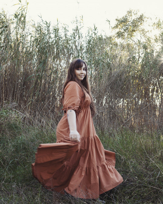 Woman wearing the Ibi Dress sewing pattern from Pauline Alice on The Fold Line. A maxi dress pattern made in cotton poplin, linen, corduroy, chambray, twill, or swiss dot fabric, featuring ties at the waist and sleeves, buttons at the back, and gathered tiers. 