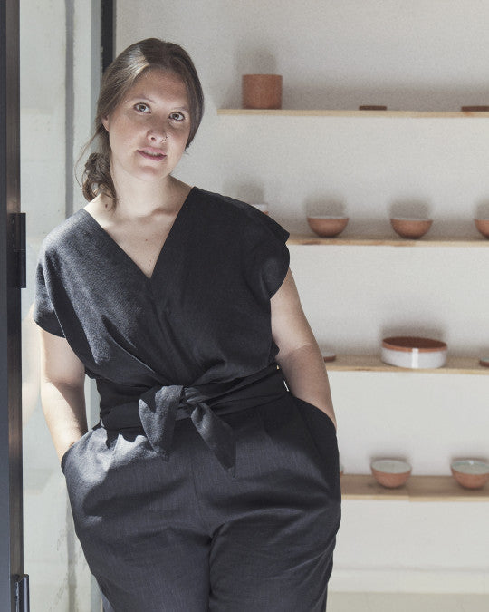 Woman wearing the Mila Jumpsuit sewing pattern from Pauline Alice on The Fold Line. A sleeveless jumpsuit pattern made in cotton, linen, silk, chambray, twill, crepe, viscose, or tencel fabric, featuring a V-neckline, high waistline, wraparound waist ties, pleated trousers, curved slanted pockets, back button and loop closure, and elasticised back waistband.