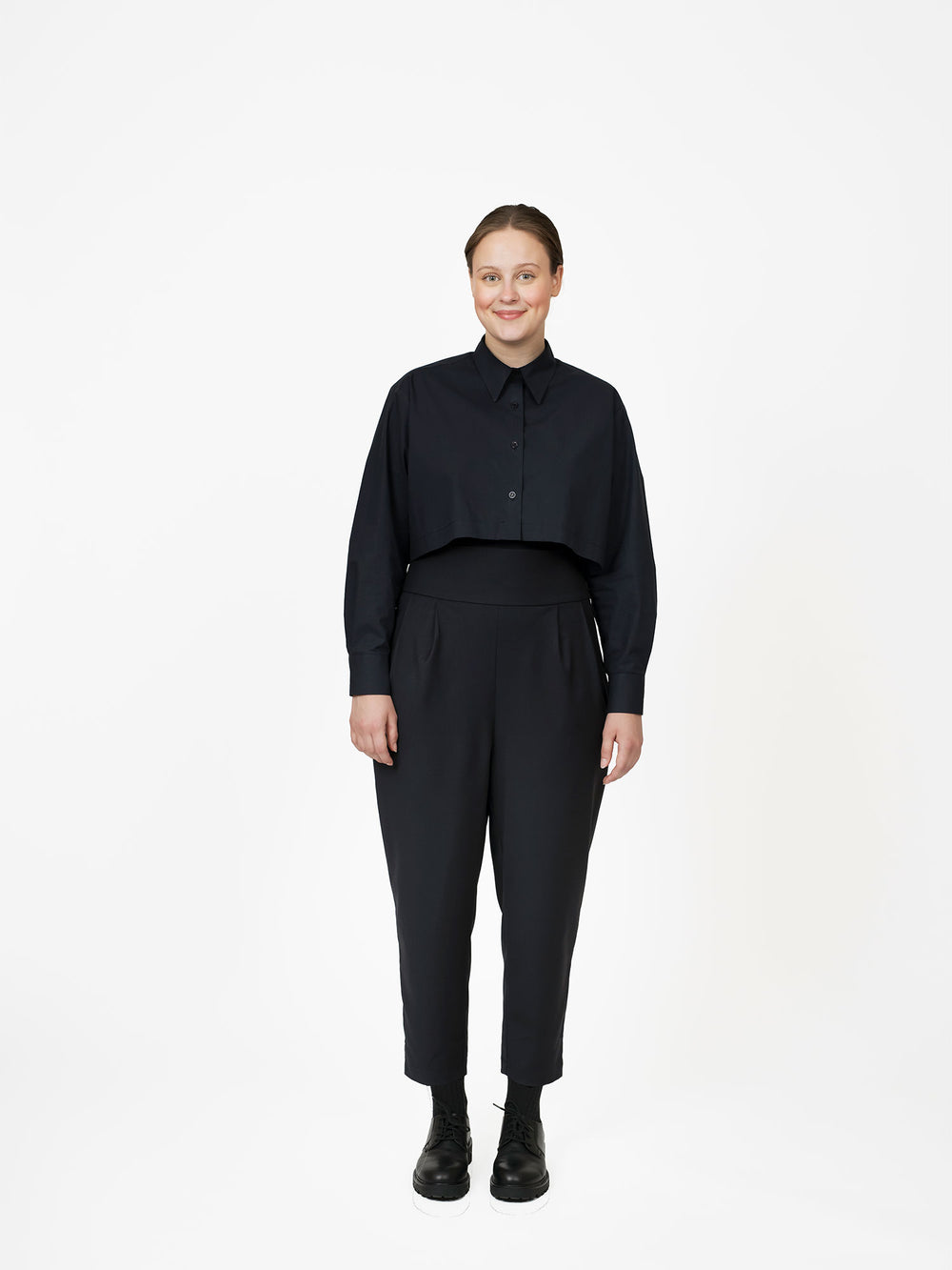 Woman wearing the Pleated Trousers sewing pattern from The Assembly Line on The Fold Line. A trousers pattern made in twill or wool fabric, featuring a high waist, dropped crotch, double pleats at the front, slanted pockets, side zipper, and cropped length. 